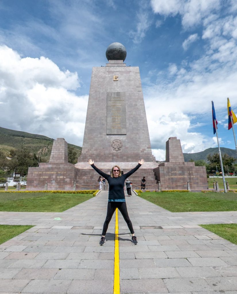 Julien Casanova, the founder of Cultures Traveled travel blog, stands with feet on either side of the painted yellow equator line at the iconic Mitad del Mundo monument.