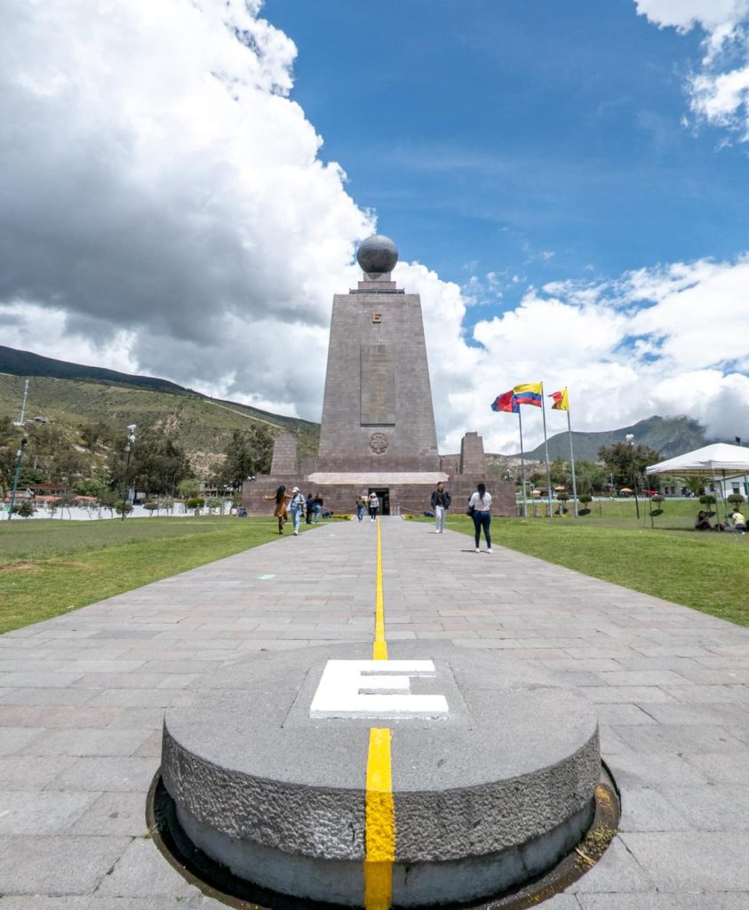 The tall Mitad del Mundo Ecuador monument with the yellow equator line stretching down the walkway, flanked by flags and green grass. In the foreground is a large E, marking the East side of the equator line in Ecuador.