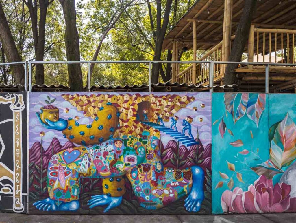 This Escher style street art depicts a body holding a planter in the shape of a face with a small plant. The body is made of small designs and small houses with tiny people carrying some of the small houses up a set of stairs.