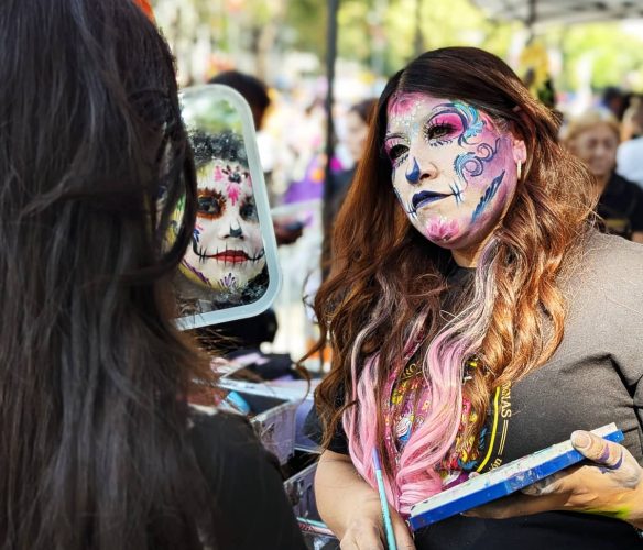 An artist applying La Catrina day of the dead makeup on a woman who checks her sugar skull design in a small mirror. The artists' own face is also painted using white, blue, pink, and purple.