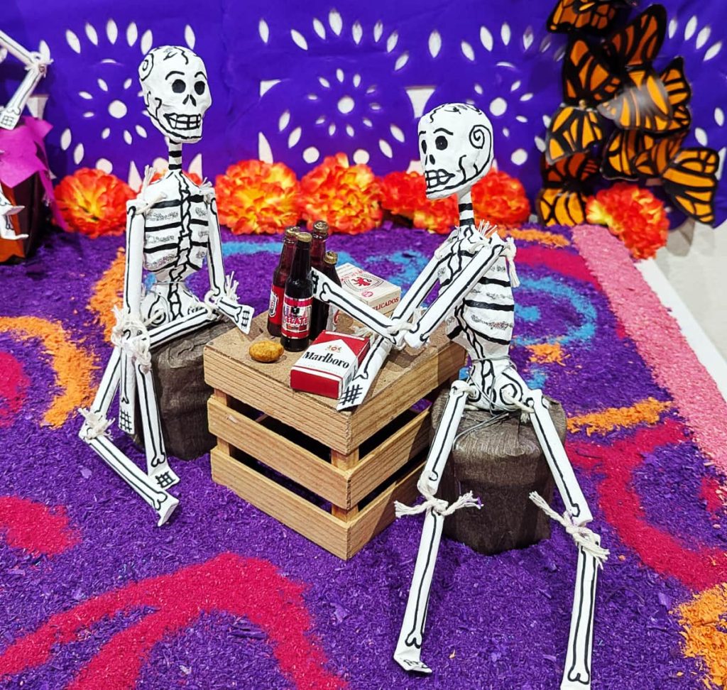 Miniature figurine, a Day of the Dead tradition, of two skeletons seated on stools around a wooden crate table with beer bottles and cigarettes with purple papel picado, butterflies, and orange marigolds.