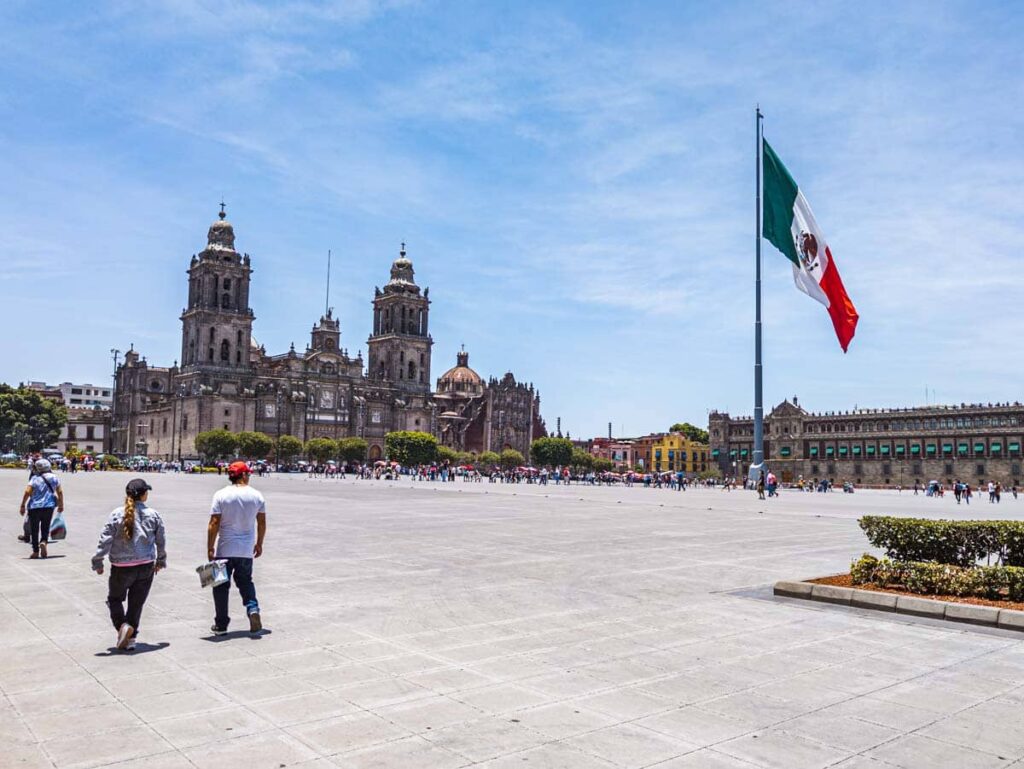 People walk through the Zocalo in Mexico City. In the large plaza the Mexican flag flies in the center, the National Palace is on the left, and the Metropolitan Cathedral is in the back.