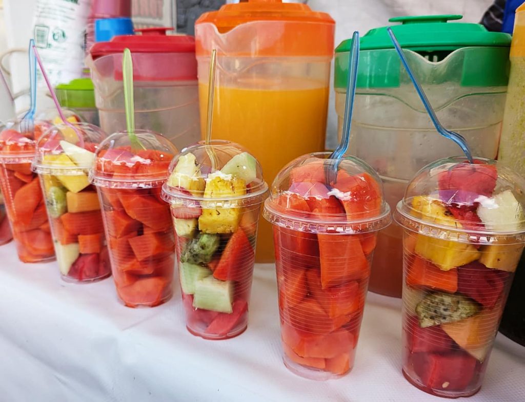 Six plastic cups filled with fresh cut tropical fruit including papaya, pineapple, and watermelon lined up at a street food cart in Mexico City, with pitchers of fresh juice in the background.
