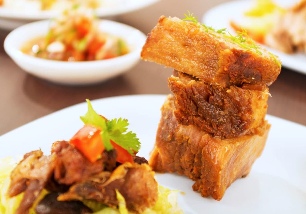 Close-up of fritada, a typical dish in Ecuador featuring crispy pieces of golden-brown pork stacked on a white plate and garnished with herbs, served with a side salad.