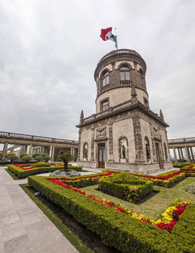 Exterior view of the garden and iconic building topped with a Mexican flag on the terrace of Chapultepec Castle in Mexico City, showcasing its beautiful garden surroundings.