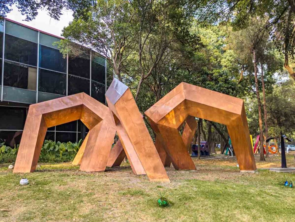 A modern metal sculpture in the garden of the Museo de Arte Moderno in Mexico City surrounded by trees near the museum building.