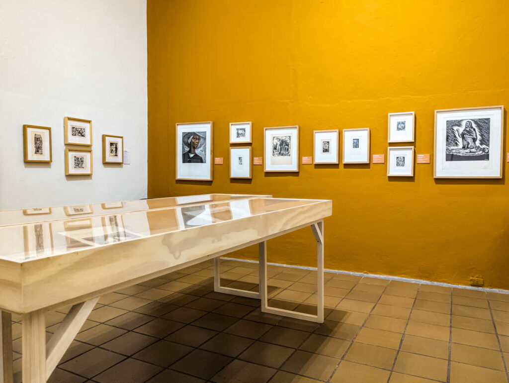 Multiple artworks hanging on the wall in a yellow room near the glass table display at the Graphic Art Museum in Mexico City.