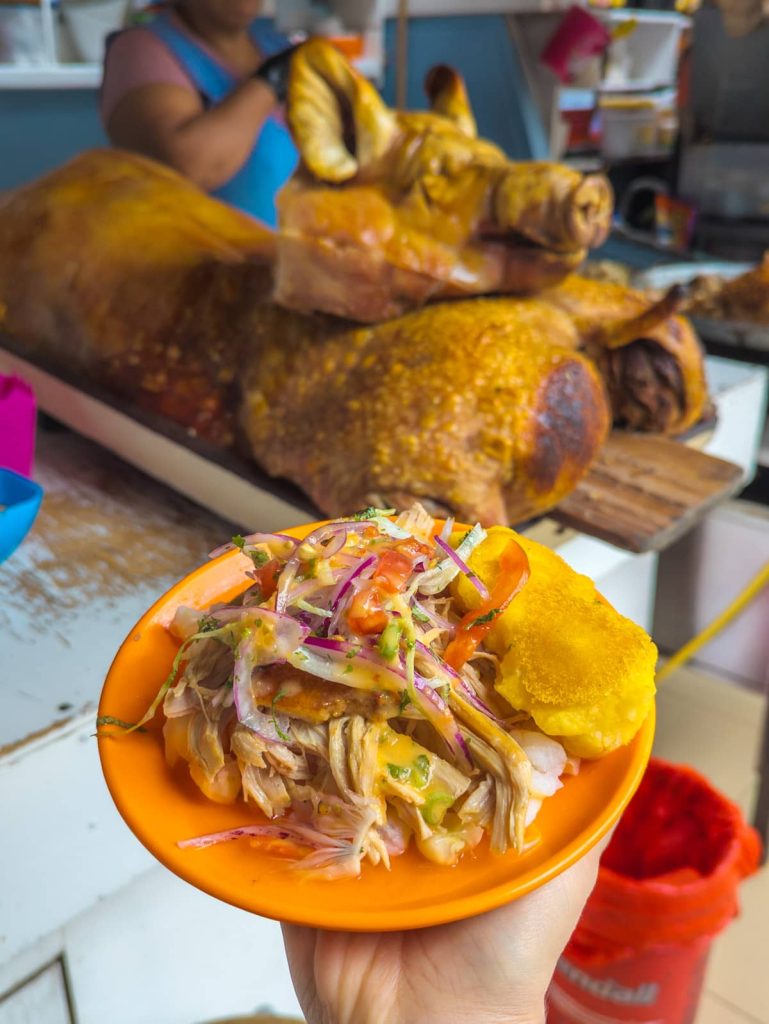 Traditional Ecuadorian hornado served on an orange plate with mote, pickled onions, and fried plantain at a Cuenca market food stall.