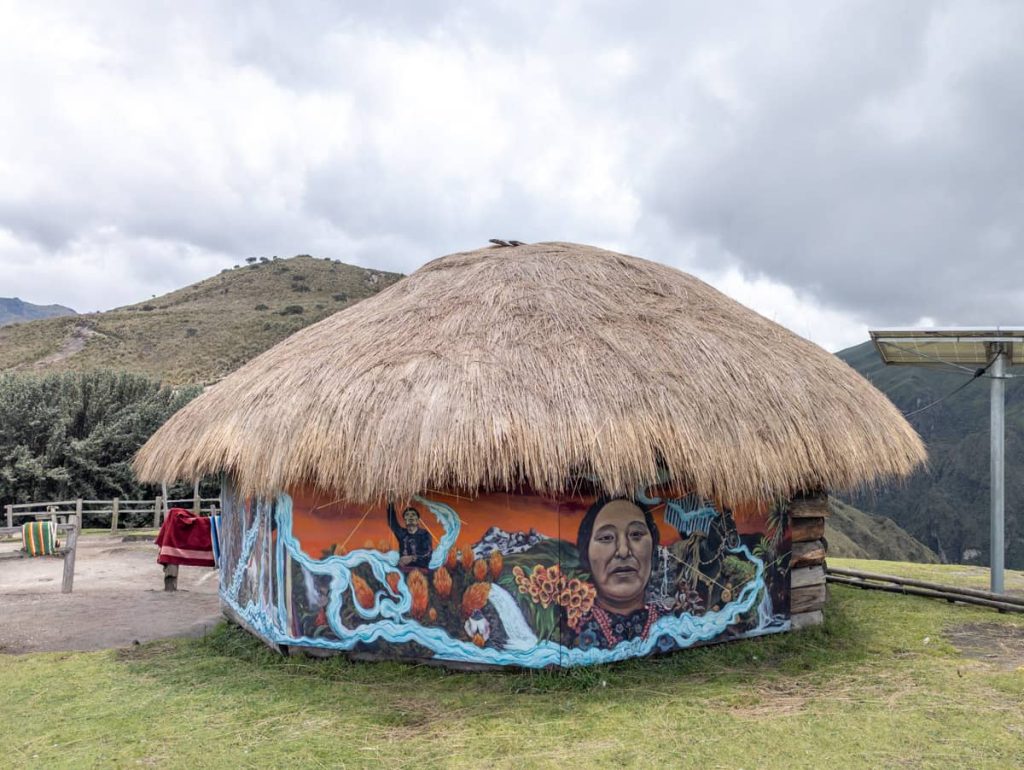 A round, thatch-roofed hut painted with colorful murals of Indigenous faces and natural scenes stands against the mountainous landscape near the cable car in Quito.