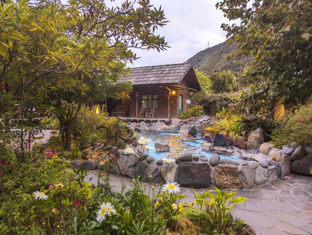 A wooden cabin hotel at Papallacta resort in Ecuador with a shallow hot spring edged with a rock wall and surrounded by plants.