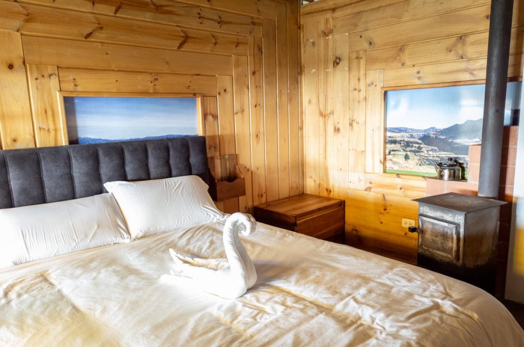 A cozy wooden cabin room at a hotel in Quilotoa Ecuador features a plush bed, towel folded into a swan, and a wood stove.