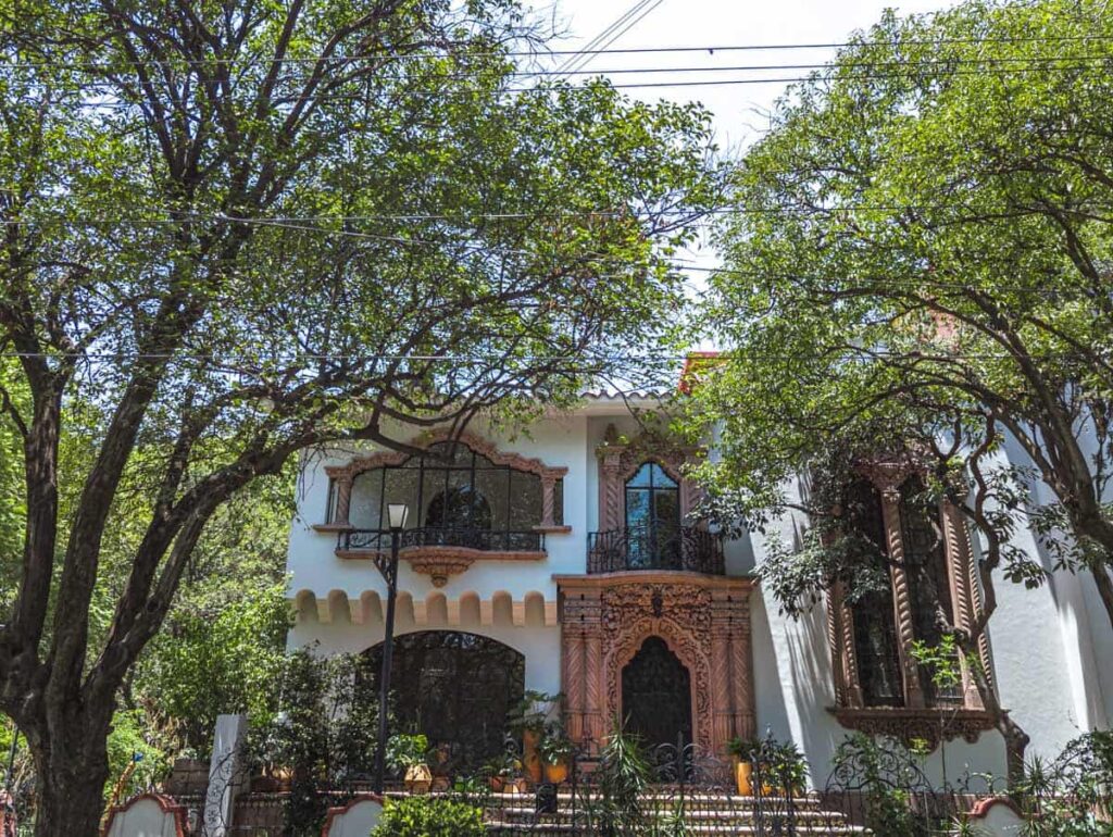 An antique white house in Polanco Mexico City showcases ornate decoration in a natural terracotta around the doors and windows with trees on either side in the foreground.