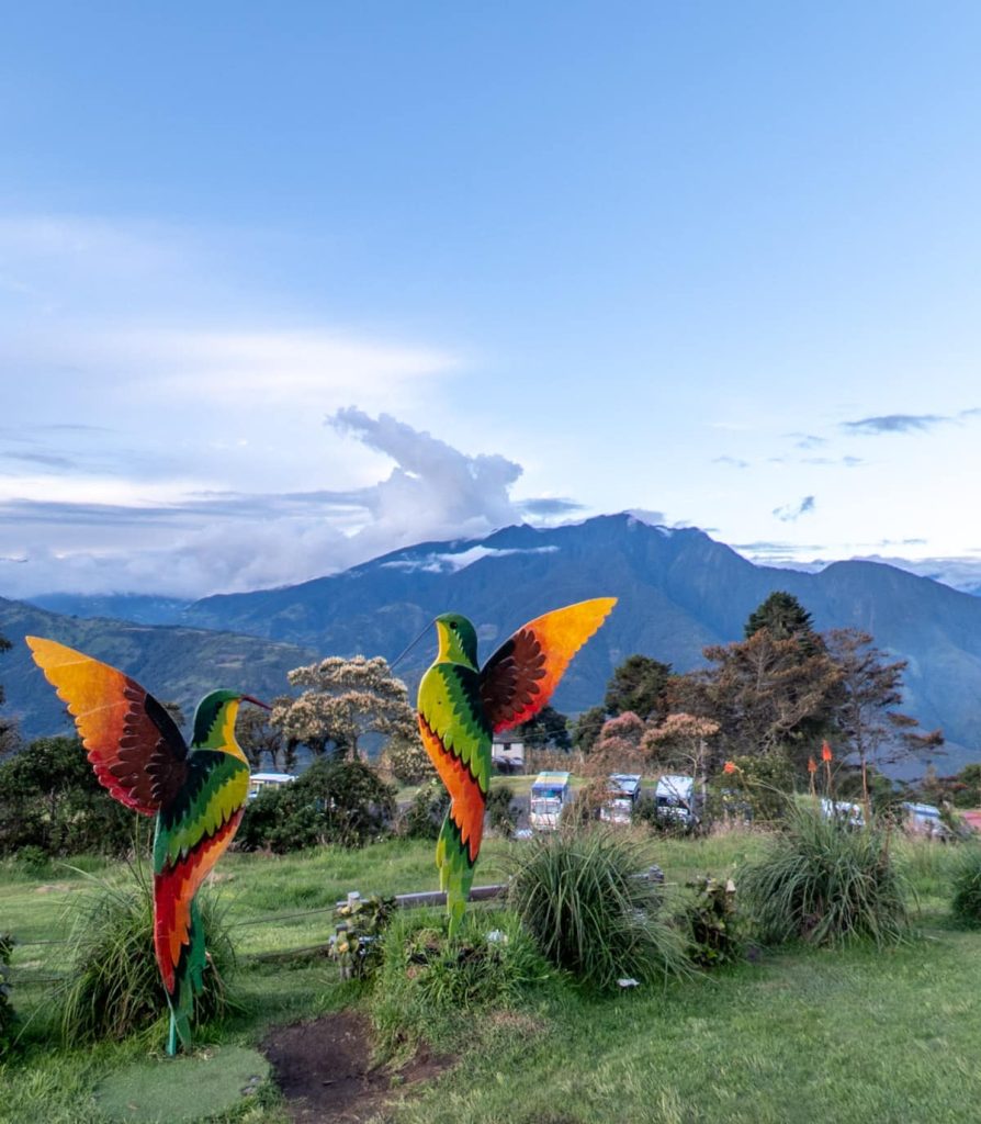 Scenic spot at Casa del Arbol and the swing at the end of the world features two vibrant hummingbird sculptures facing the mountains of Baños, Ecuador, with cotton-like clouds and layered green hills behind.