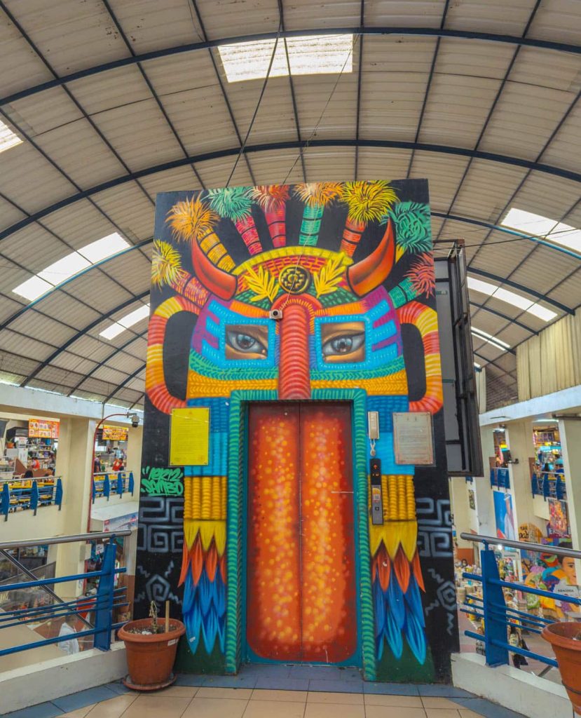 A colorfully painted elevator in Mercado 10 de Agosto Cuenca, decorated with a vibrant Inti Raymi mask mural under the market’s arched roof.