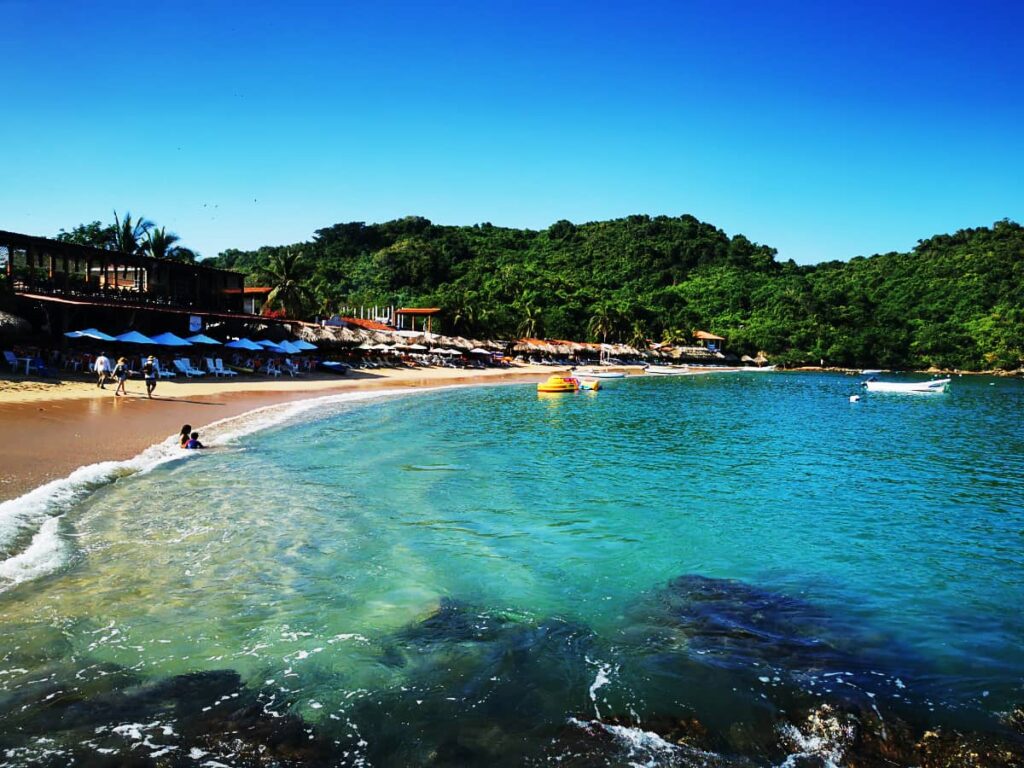 Waves gently roll to shore in this overall shot of Playa Varadero in Isla Ixtapa. Several people walk along the beach in front of the restaurants as two kids sit in the shallow waves. The water is a vibrant turquoise with verdant green trees on the hill behind.