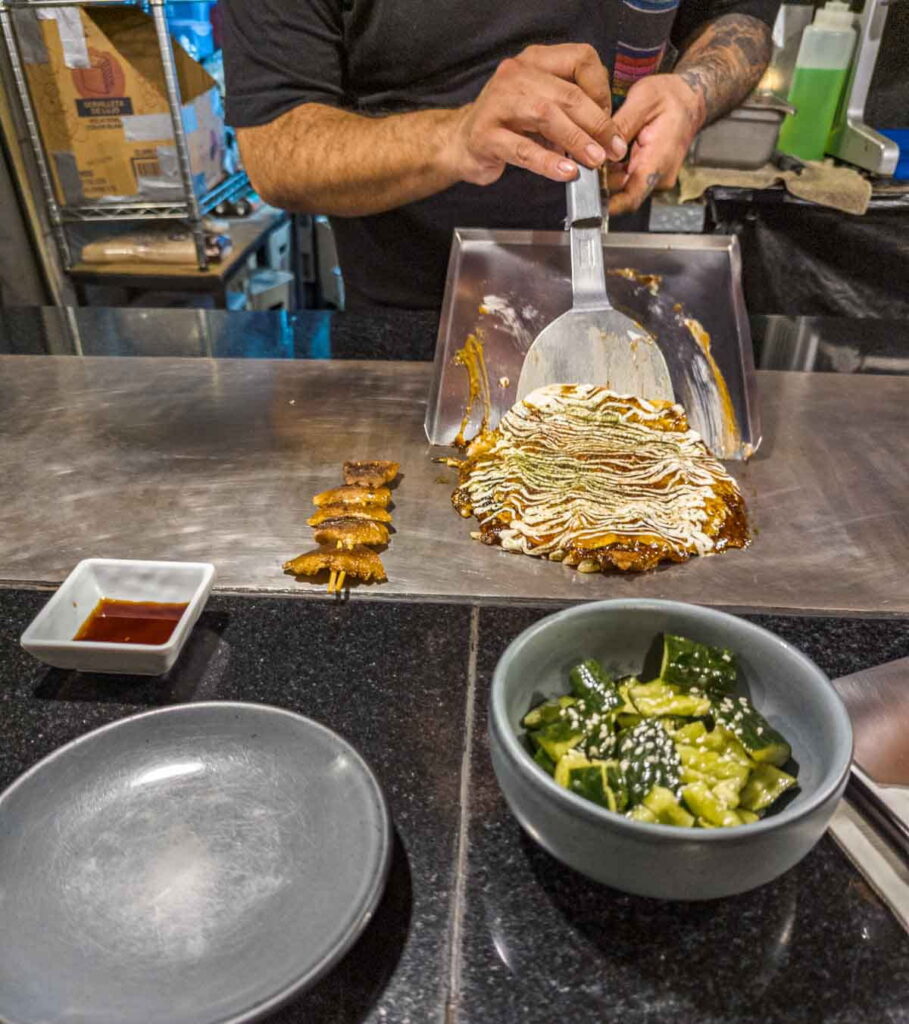 A server uses a spatula to slide okonomiyaki, a traditional Japanese food, onto a grill at a Japanese restaurant in CDMX that features street food.