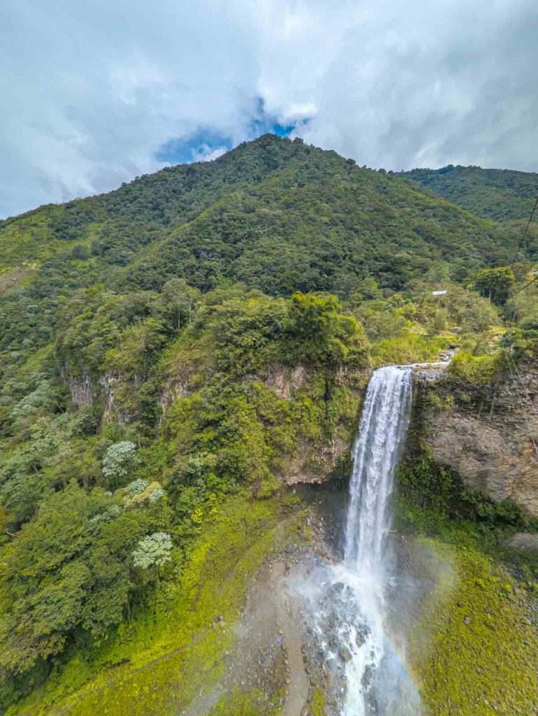 A dramatic overhead close-up view of a tall waterfall cascading down a mossy cliff into rocky terrain, captured while on a zipline during a Baños waterfall tour.