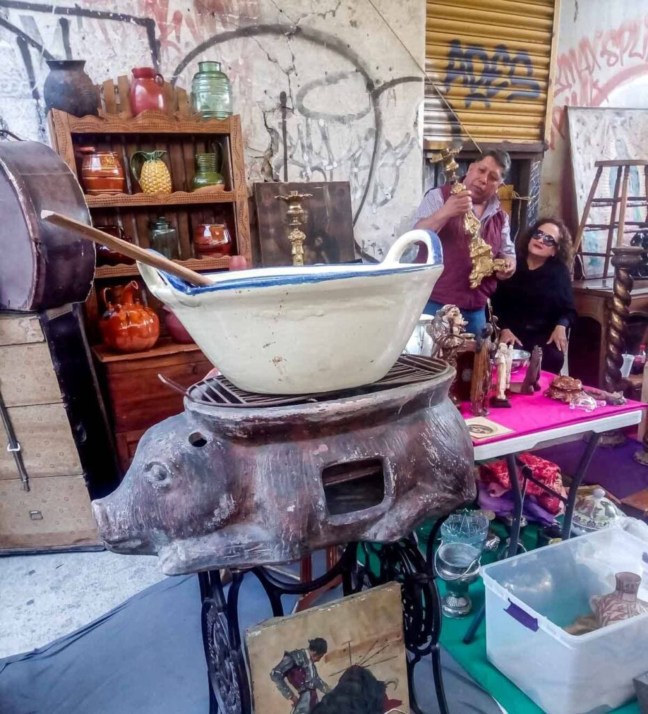 A flea market in Mexico City with different items for sale including a large grill in the shape of a pig. In the background, a couple are admiring a candle holder.