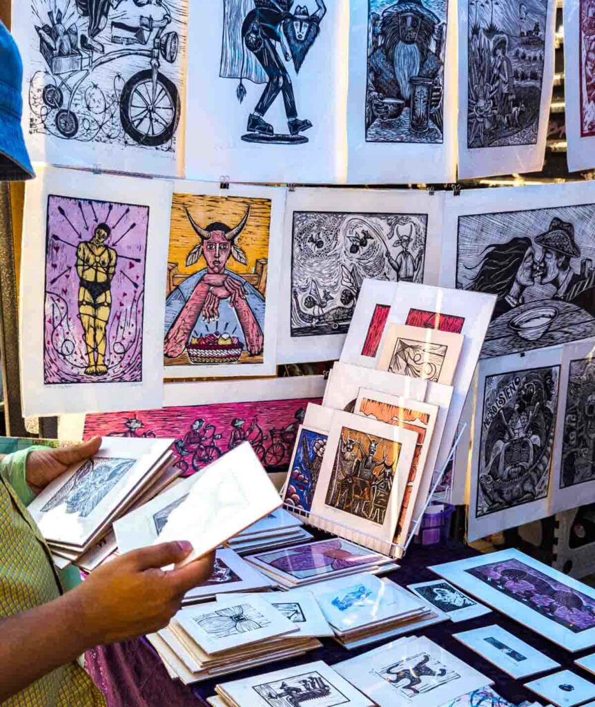 At the Lagunilla market in Mexico City, a customer holds several pieces of graphic artwork in front of a table display and hanging display.