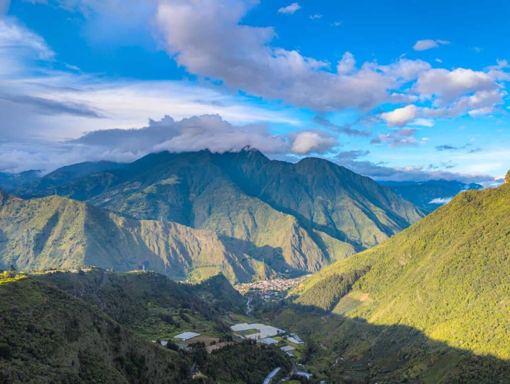 A panoramic view of Baños Ecuador shows a small town nestled between steep green mountains with scattered farms and clouds rolling over the peaks in the distance.
