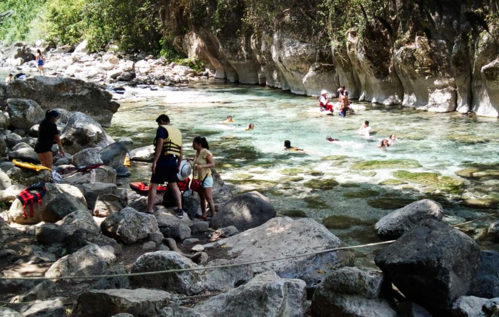 People swim in the clear blue river at Puente de Dios, Huasteca Potosina. In the foreground are large rocks with several people wearing life jackets.