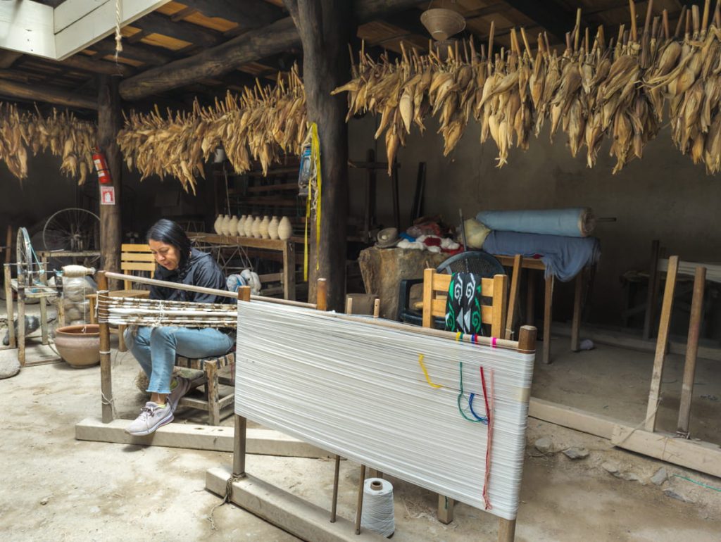During an artisan day tour from Cuenca, a woman works with yarn in a traditional weaving workshop, with corn husks hanging overhead.