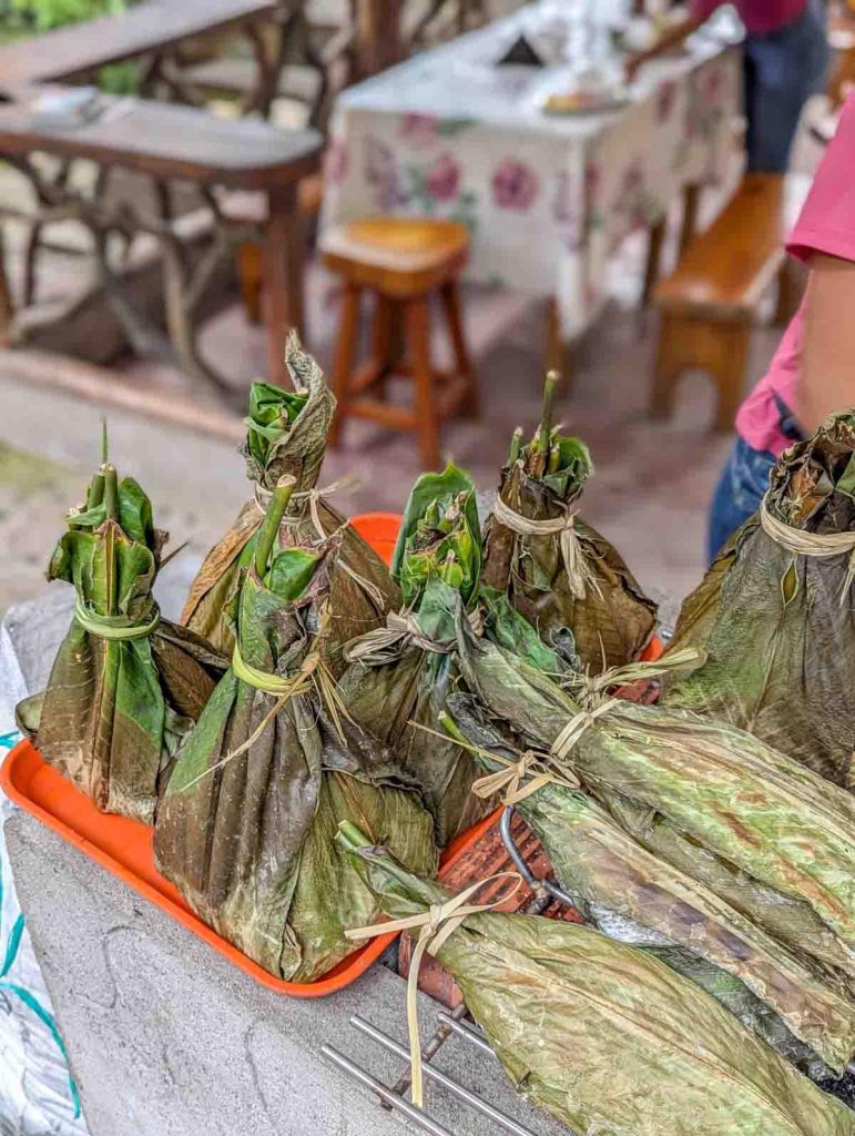Several maitos wrapped in green bijao leaves and tied with natural fiber, displayed on a red tray at an outdoor food stand. Maito is a traditional Amazonian dish in Ecuador, often filled with fish or chicken and grilled over hot coals.