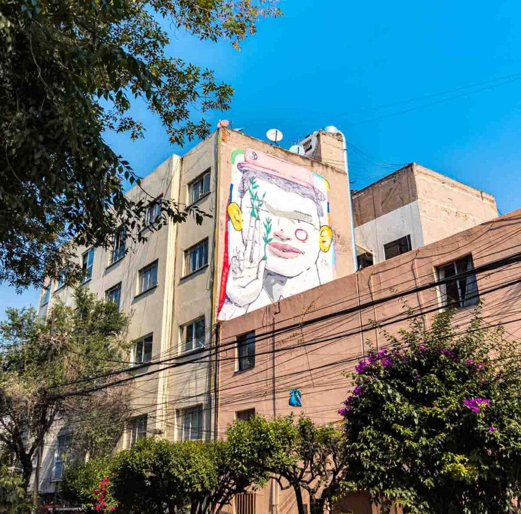 A man's face with his hand posing a "peace sign" is painted on the side of a buildings in this pastel color street art in Mexico City. His pink hat features a small yin-yang symbol.