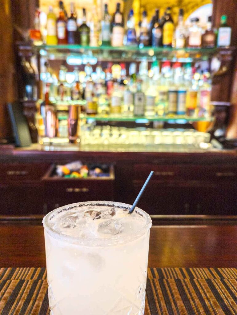 A margarita on the rocks with a salt rim sits on the bar at La Opera, a Mexico City cantina with an infamous past. At the back of the bar are backlit shelves of liquor bottles.
