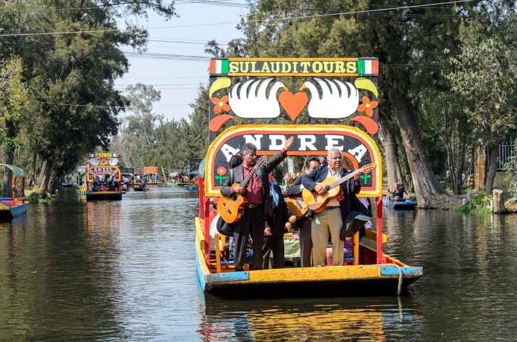 Mariachi band singing performance at a colorful boat called Andrea in Mexico City's canal with other trajineras in the back.