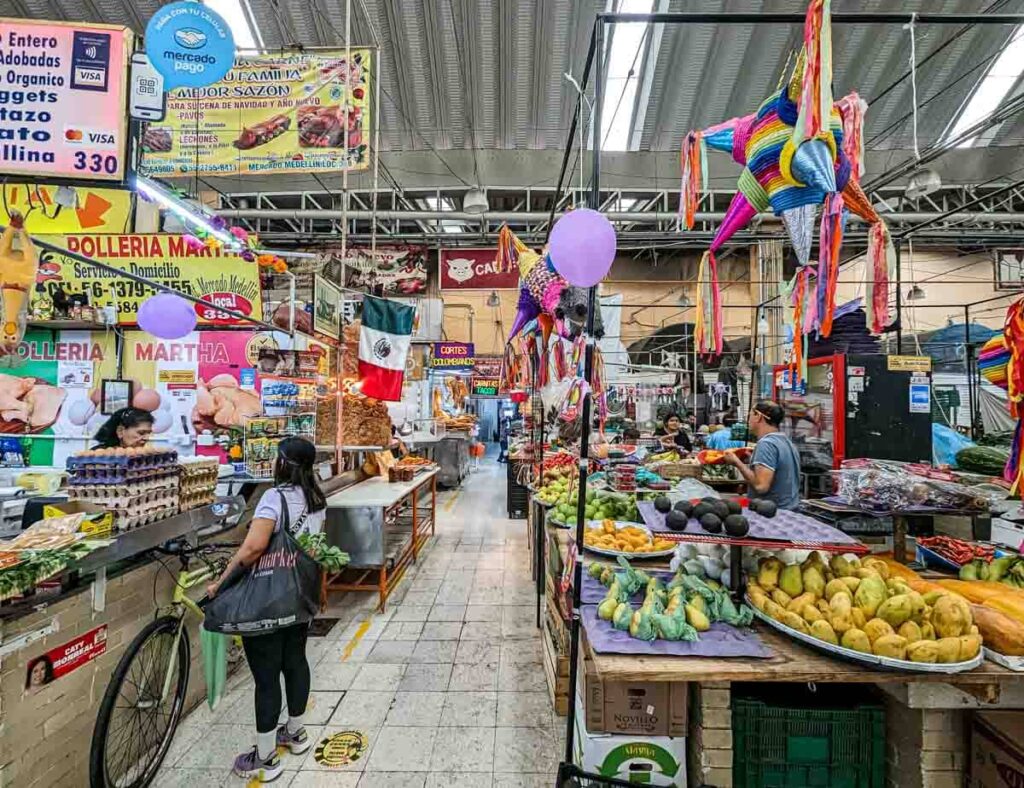 Fruit vendors with colorful pinata decorations at Medellin Market in Mexico City. A customer shops at one of the stands while holding a bag full of products and her bicycle.