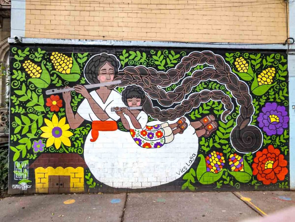 This street art in CDMX depicts a mother and child with long braids playing flutes surrounded by various flowers and ears of corn.