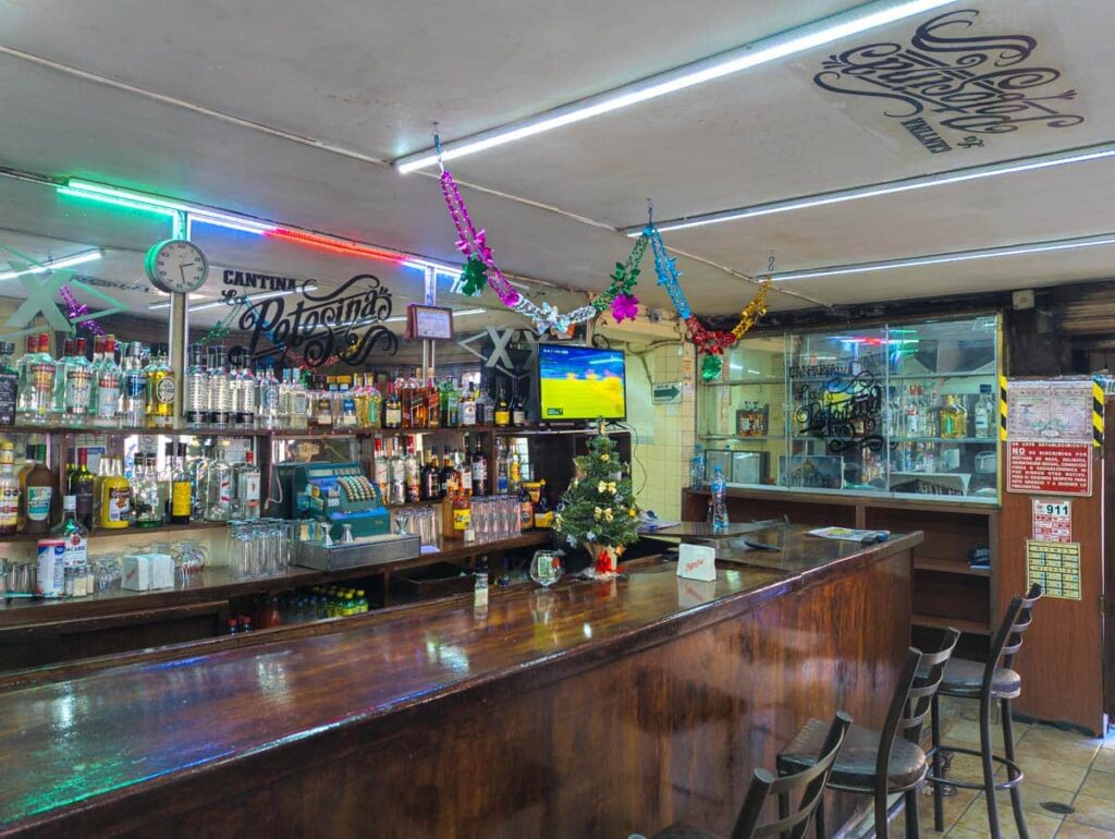 The bar at Mexico City cantina La Potosina features liquor bottles, and old cash register, and a TV playing a soccer match. In front of the bar are a few empty bar stools.