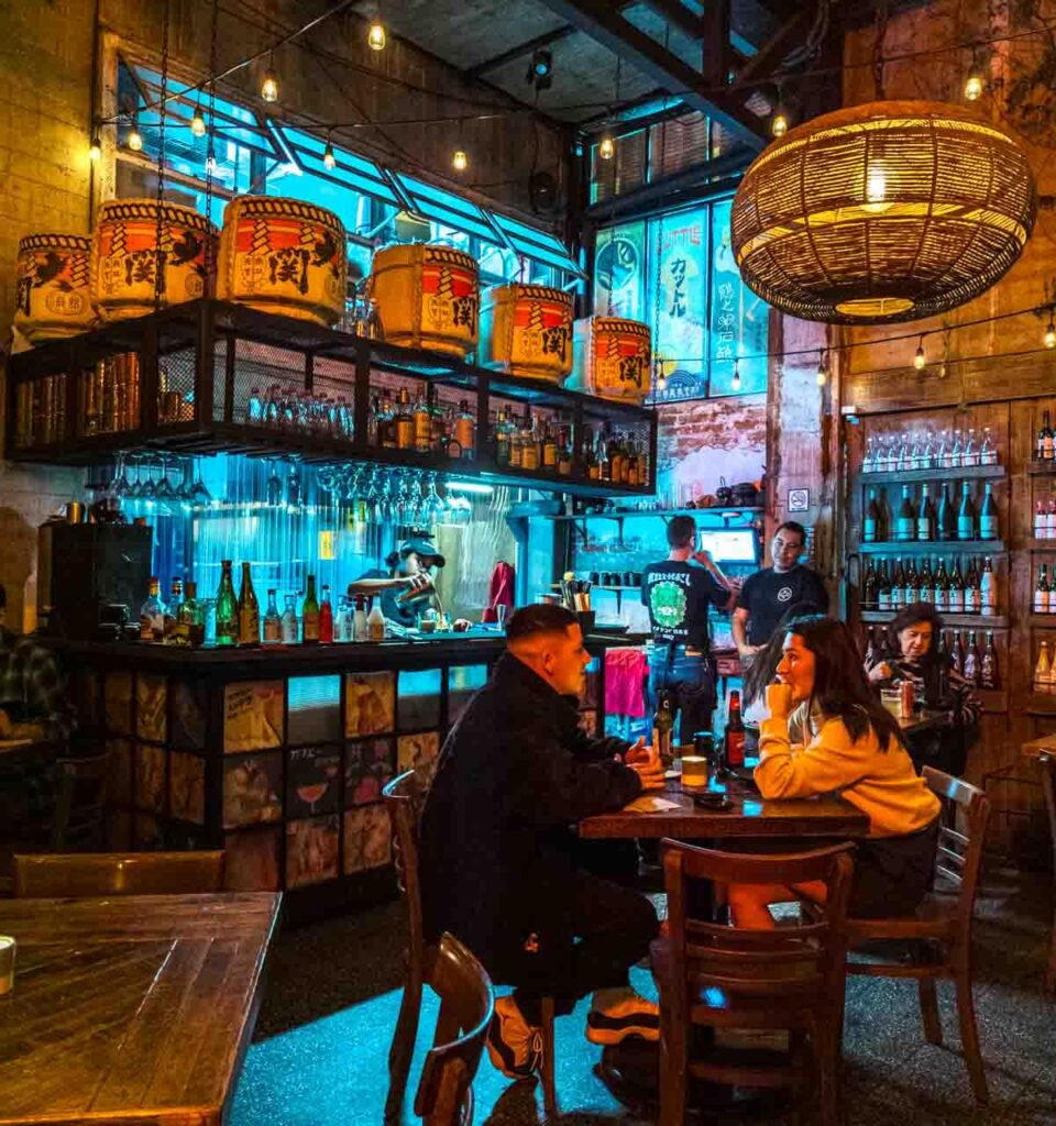 A couple sits at a table at Koku Japanese restaurant in Mexico City. Behind them the bar features large containers with Japanese print and a few staff working.