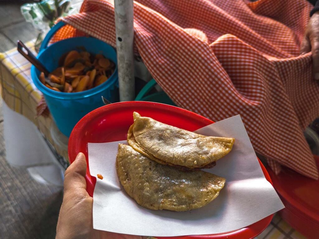 Two tacos de canasta in Mexico City on a red plate lined with a thin paper. In the background the basket of tacos is covered with a red an white cloth. A large blue tub of pickled carrots and jalapeno is on the left.