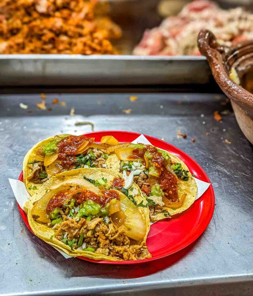 Three tacos on a red plate sit on a metal serving bar at El Gato Volador, a Mexico City taqueria in Roma Norte. In the background you can see the grill with piles of meat cooking.