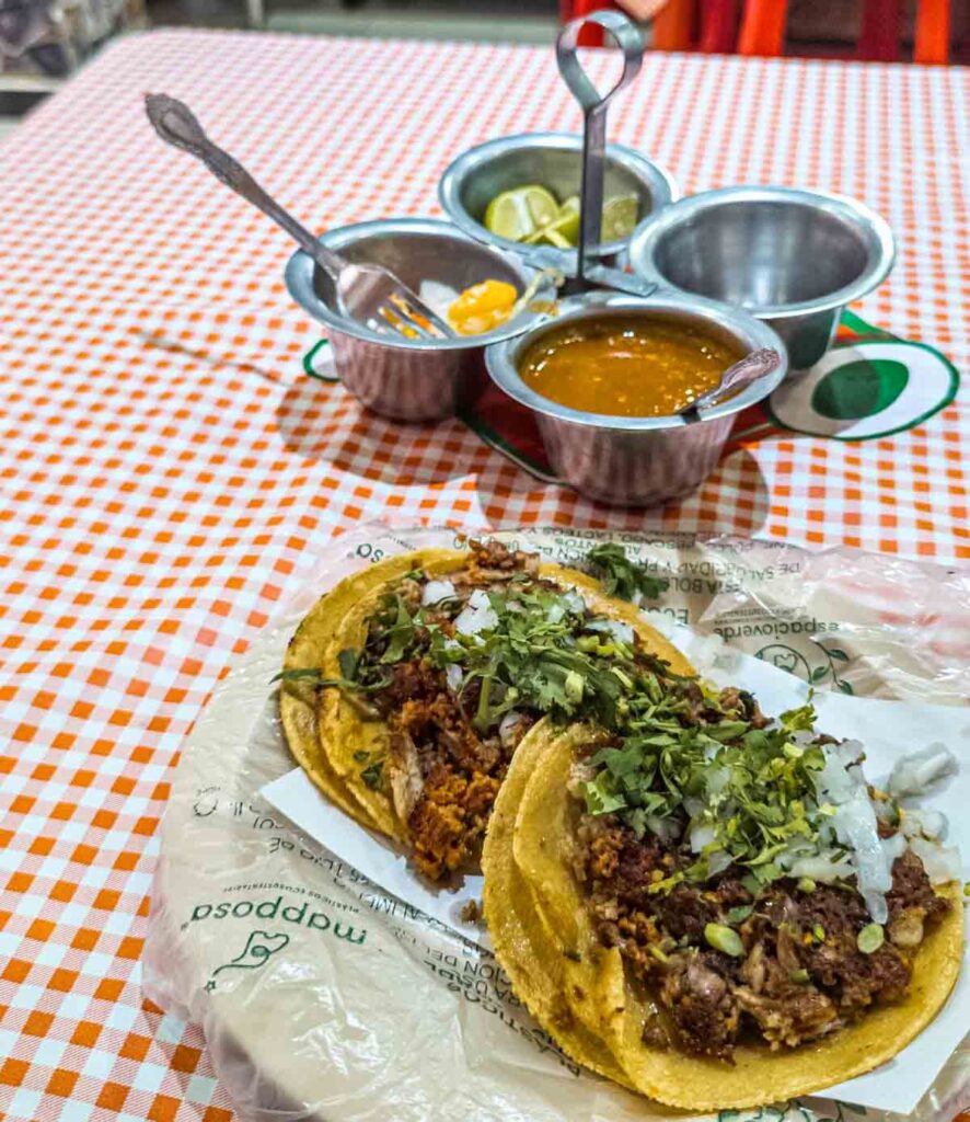 Two tacos served with double tortillas and topped with onion and cilantro sit on a plate on a checkered table cloth. In the background is a small metal caddy that hold different salsas and limes.
