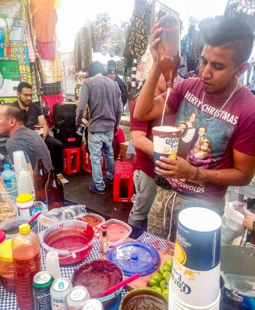 At the Lagunilla Sunday market, a man pours a large bottle of beer into a cup lined with a red chili syrup to make a michelada. In front of him are containers of spices and other syrups.