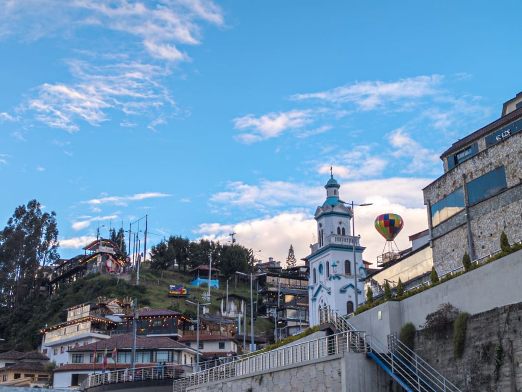 Colorful hillside view of Turi, a popular viewpoint in Cuenca Ecuador, with a white church tower, a hot air balloon structure, and restaurants decorated with string lights. Visiting Turi is a top recommendation for what to do in Cuenca, offering panoramic views and local charm.