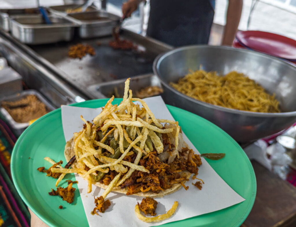 A green plate of mixiote tacos in Mexico City are topped with shoestring potatoes. In the background is a large metal bowl of the potatoes and a flat top grill.