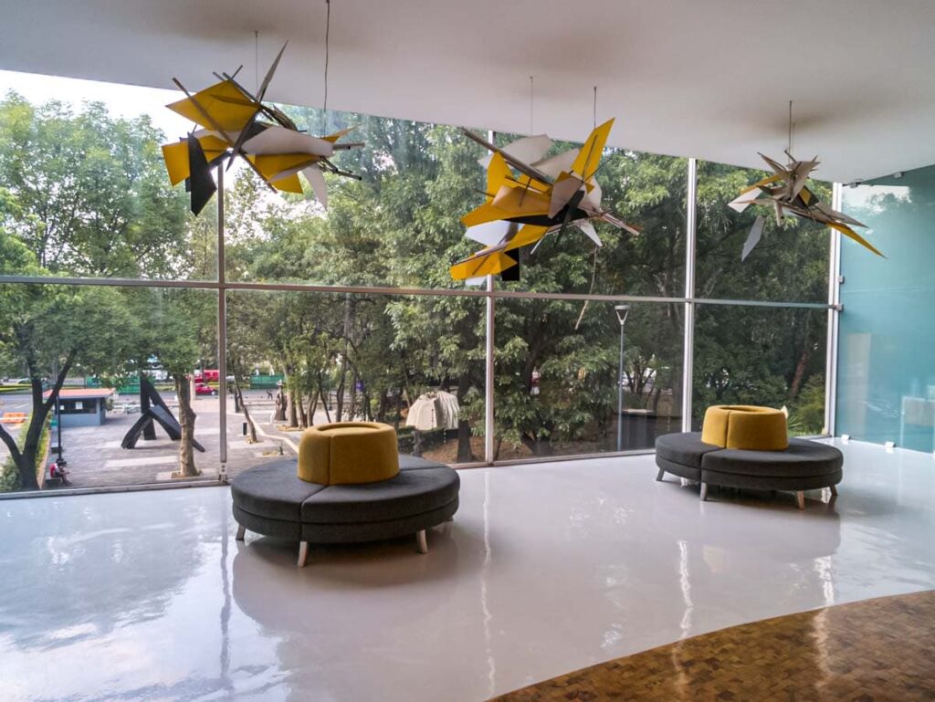 Two black and yellow round chairs below the abstract design hanging from the ceiling next to the glass windows at the Modern Art Museum in Mexico City.