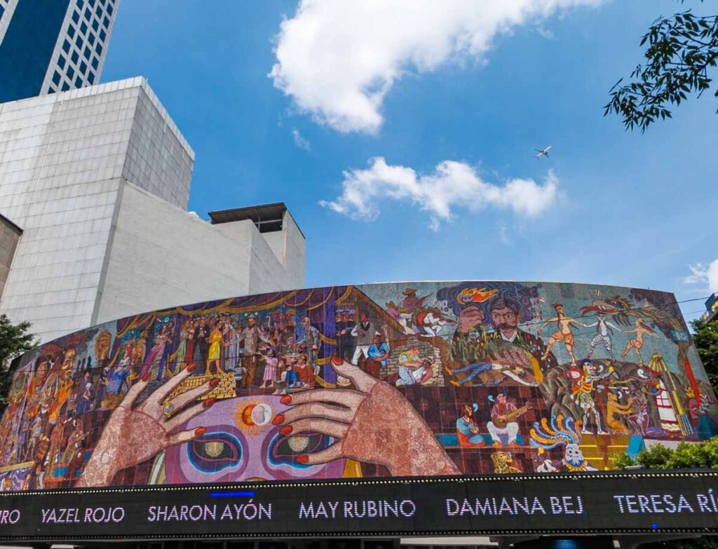 The exterior of the Insurgentes Theater in CDMX featuring a Diego Rivera mosaic under a blue sky with white clouds and an airplane. The main figure in the mural is a large masked face with hands painted with red nail polish partially in front of the mask.