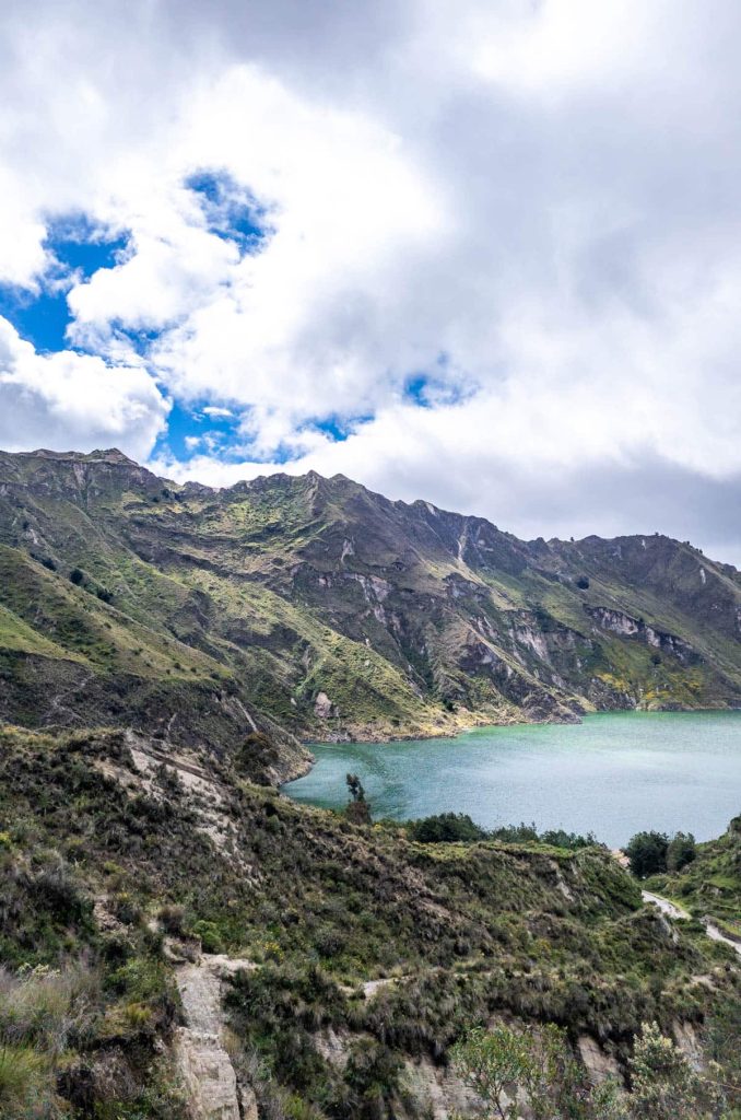 A view of green mountains surrounding the turquoise water of Quilotoa Lake beneath a mostly cloudy sky. This viewpoint is a highlight of visiting Quilotoa from Quito.