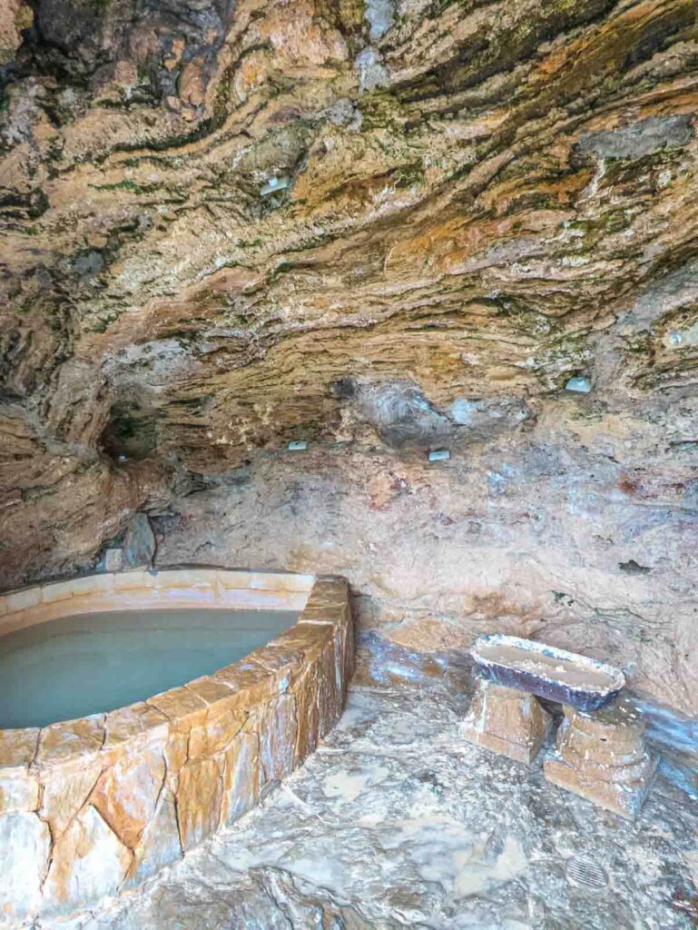 Private stone soaking pool inside a natural cave at Novaqua thermal baths, surrounded by mineral-streaked rock walls and a rustic stone bench.