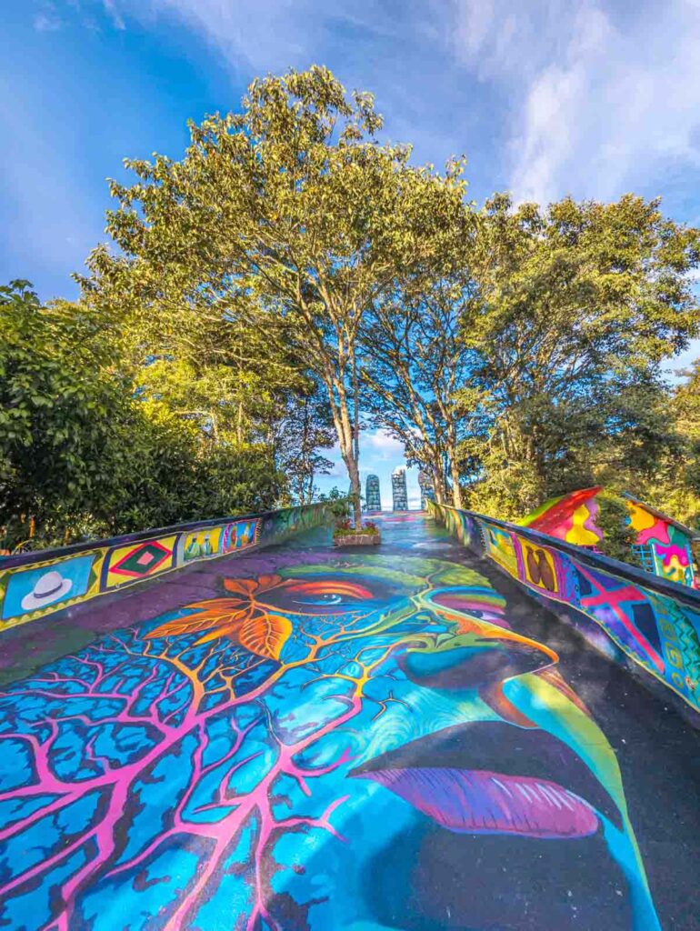 A colorful mural of a face with tree-like veins spans the walkway leading to La Mano de la Pachamama in Baños Ecuador. Trees line the vibrant path on both sides.