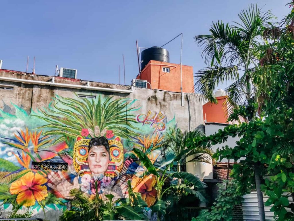 A tropical mural in Zihuatanejo Mexico painted on the side of a building depicts a woman's face with the background of a jaguar mask surrounded by tropical plants.