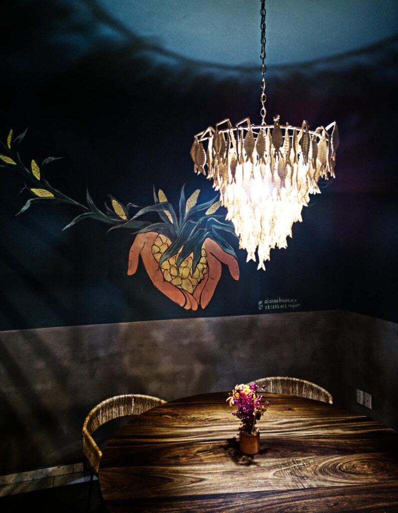 Interior of the Ojo de Agua, a vegan restaurant in Mexico City, features a wall mural of a corn plant with a pair of cupped hands holding corn kernels. In front of the wall is a wooden table with dramatic lighting.