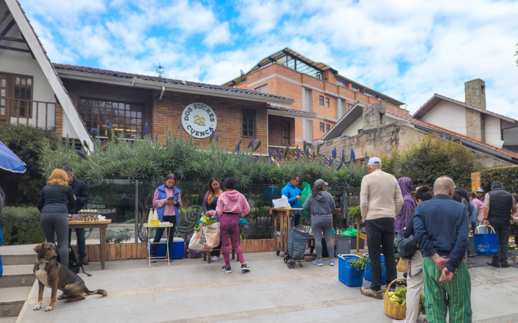 People shopping outside at the Dos Sucres organic farmers market in Cuenca. The setting is a rustic brick building in a modern neighborhood setting.