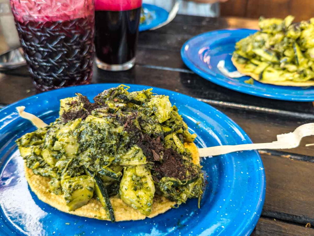 Vegan aguachile made with oyster mushrooms and cucumbers in a spicy green sauce is topped on a crispy tostada and served on a blue plate at the Lagunilla Flea Market in Mexico City. In the background, deep red cocktails are in glasses.