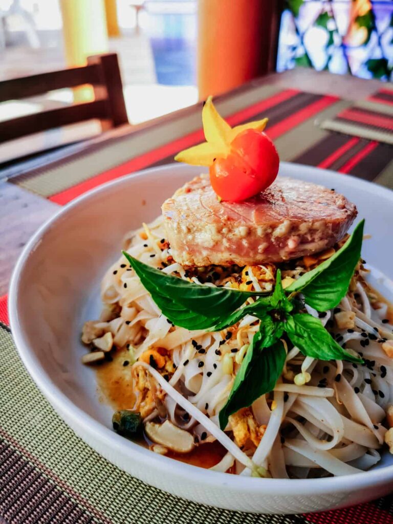 A vibrant bowl of Pad Thai topped with a seared tuna steak, fresh basil, and a decorative star fruit slice, served at a restaurant in Zihuatanejo.